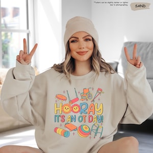 Peut inclure: Un sweat-shirt beige clair avec un motif graphique d'outils d'ergoth&eacute;rapie et le texte "HOORAY IT'S AN OT DAY!" en lettres color&eacute;es.