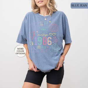 May include: Blue jean colored t-shirt with a vintage 1986 graphic. The graphic includes a cassette tape, lightning bolt, and a disco ball. The shirt has short sleeves and a relaxed fit.