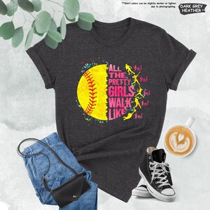 May include: Dark grey heather t-shirt with a softball graphic. The graphic features a yellow softball with red stitching and the text "ALL THE PRETTY GIRLS WALK LIKE THIS!" in pink and white. The shirt is paired with blue jeans, a black purse, and black high-top sneakers.