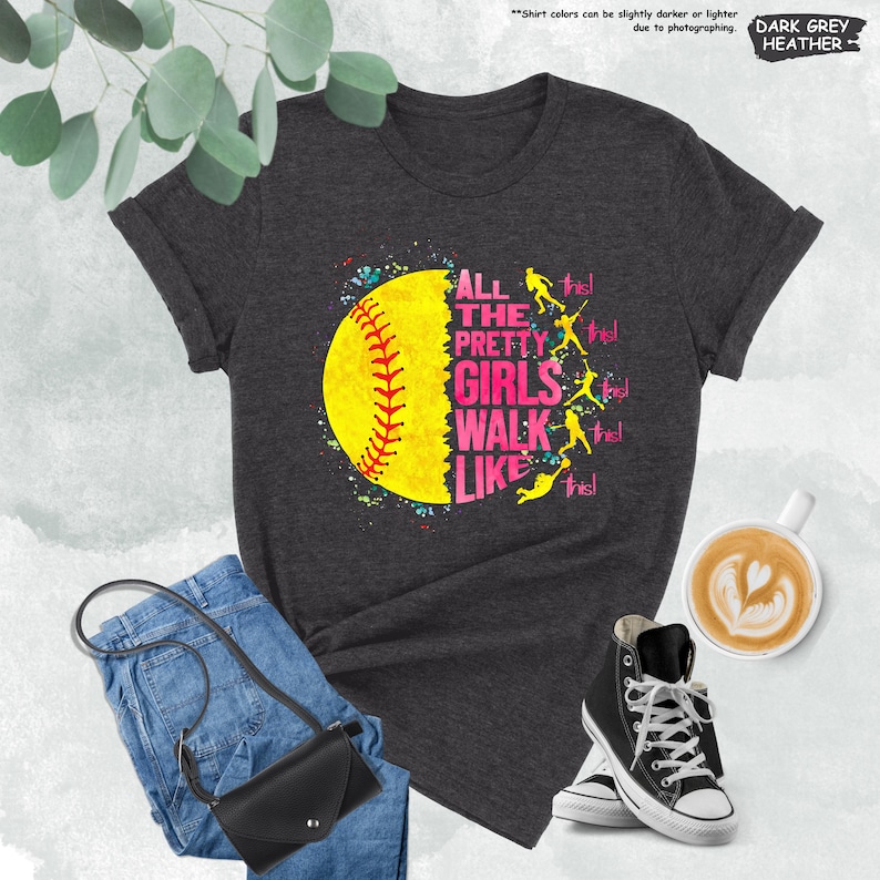 May include: Dark grey heather t-shirt with a softball graphic. The graphic features a yellow softball with red stitching and the text "ALL THE PRETTY GIRLS WALK LIKE THIS!" in pink and white. The shirt is paired with blue jeans, a black purse, and black high-top sneakers.