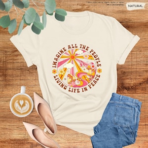 May include: A white t-shirt with a retro-style graphic design featuring a sun, clouds, mushrooms, and the text "Imagine All The People Living Life In Peace".