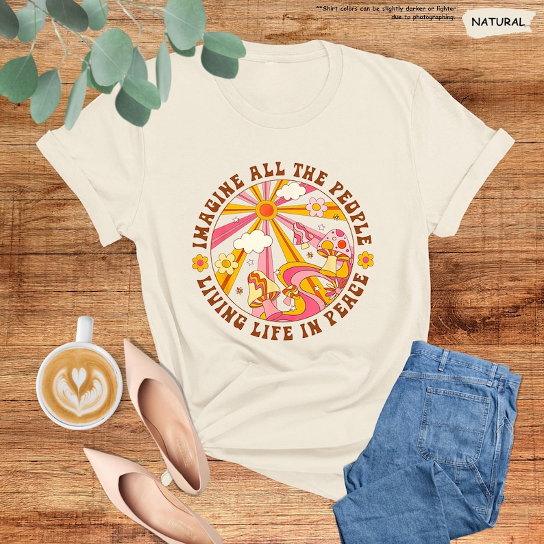 May include: A white t-shirt with a retro-style graphic design featuring a sun, clouds, mushrooms, and the text "Imagine All The People Living Life In Peace".