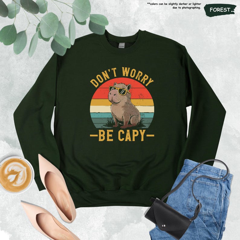 May include: Dark green sweatshirt with the text "DON'T WORRY - BE CAPY-" and a graphic of a capybara wearing sunglasses. The design is set against a retro sunset background. The sweatshirt is a forest green color.