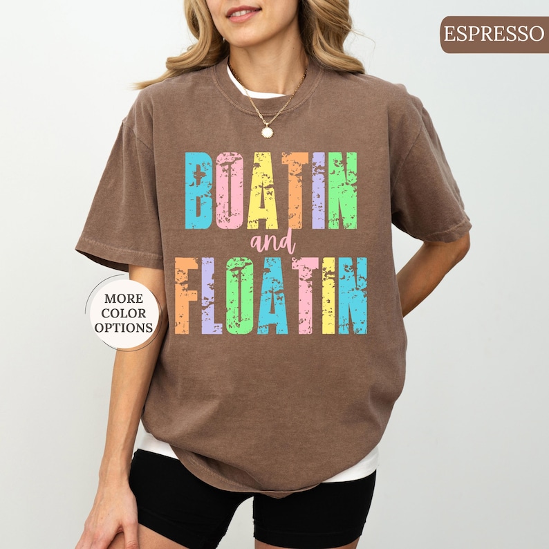 May include: A brown t-shirt with the words "BOATIN and FLOATIN" in colourful, distressed lettering. The shirt has short sleeves and a relaxed fit. A button on the left side says "MORE COLOR OPTIONS".