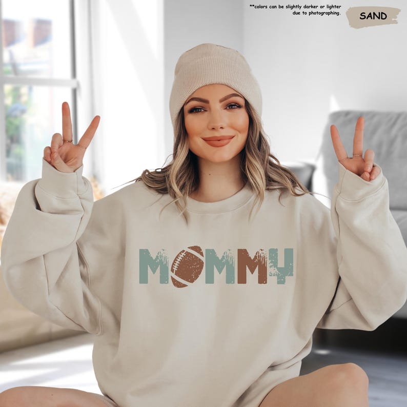 May include: A beige sweatshirt with the word "MOMMY" in distressed blue and brown letters with a brown football in the middle of the word.