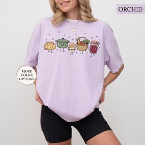 May include: An orchid-colored t-shirt featuring a graphic print of a smiling loaf of bread, a green cooking pot, an egg in an apron, a basket of vegetables, and a jar of jam, all with small stars and hearts scattered above them.