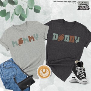 May include: Two t-shirts with a distressed vintage look. The grey shirt says "MOMMY" with a football graphic. The dark grey shirt says "DADDY" with a football graphic.