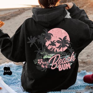 May include: Black hoodie featuring a beach-themed design on the back. The design includes palm trees, a pink sun, and the words "Beach Bum". The hoodie is worn on a sandy beach. The text "Beach Bum" is in a script font.