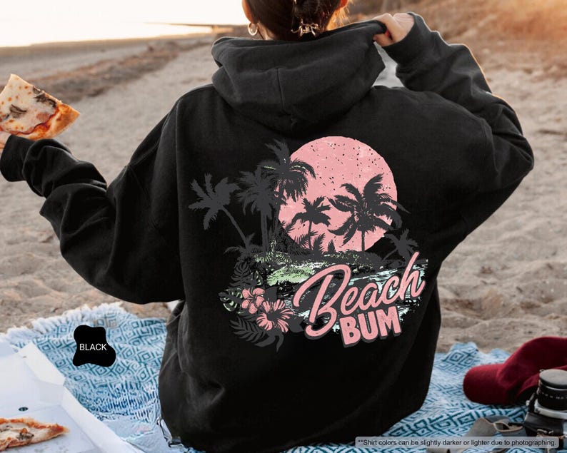 May include: Black hoodie featuring a beach-themed design on the back. The design includes palm trees, a pink sun, and the words "Beach Bum". The hoodie is worn on a sandy beach. The text "Beach Bum" is in a script font.