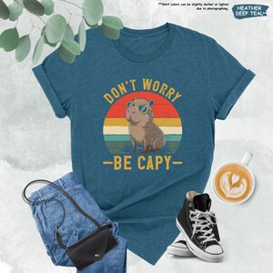 May include: Teal t-shirt with the text "DON'T WORRY - BE CAPY" and a graphic of a capybara wearing sunglasses against a sunset background. The shirt is paired with blue jeans, a black purse, and black high-top sneakers.