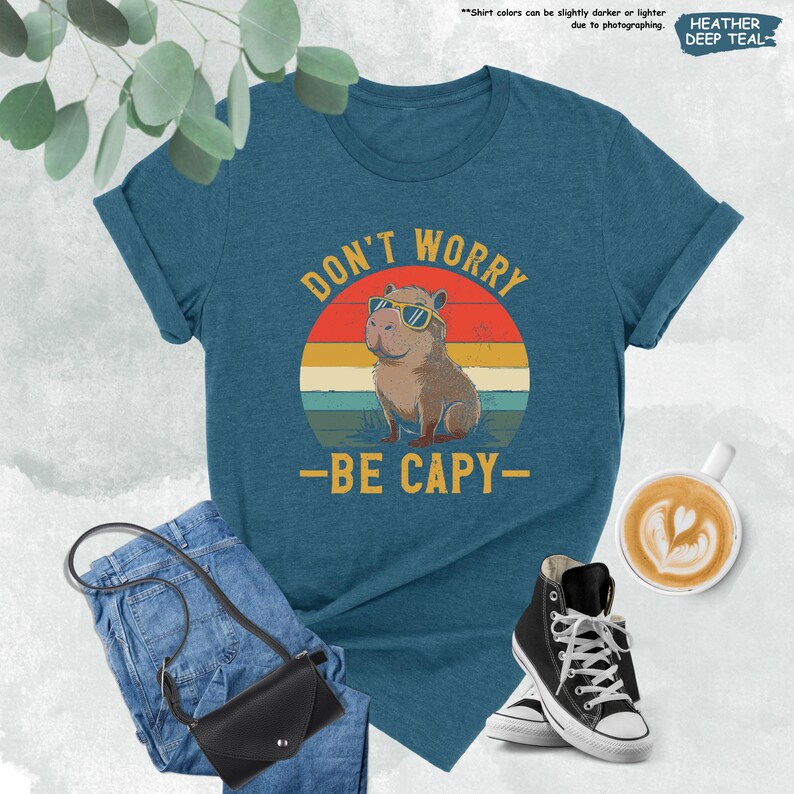 May include: Teal t-shirt with the text "DON'T WORRY - BE CAPY" and a graphic of a capybara wearing sunglasses against a sunset background. The shirt is paired with blue jeans, a black purse, and black high-top sneakers.