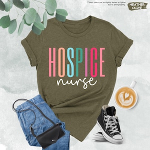K&ouml;nnte beinhalten: Ein gr&uuml;nes T-Shirt mit dem Text "HOSPICE nurse" in bunten Buchstaben. Das Shirt liegt auf einer wei&szlig;en Oberfl&auml;che mit einer Jeans, einer Handtasche und einer Tasse Kaffee.