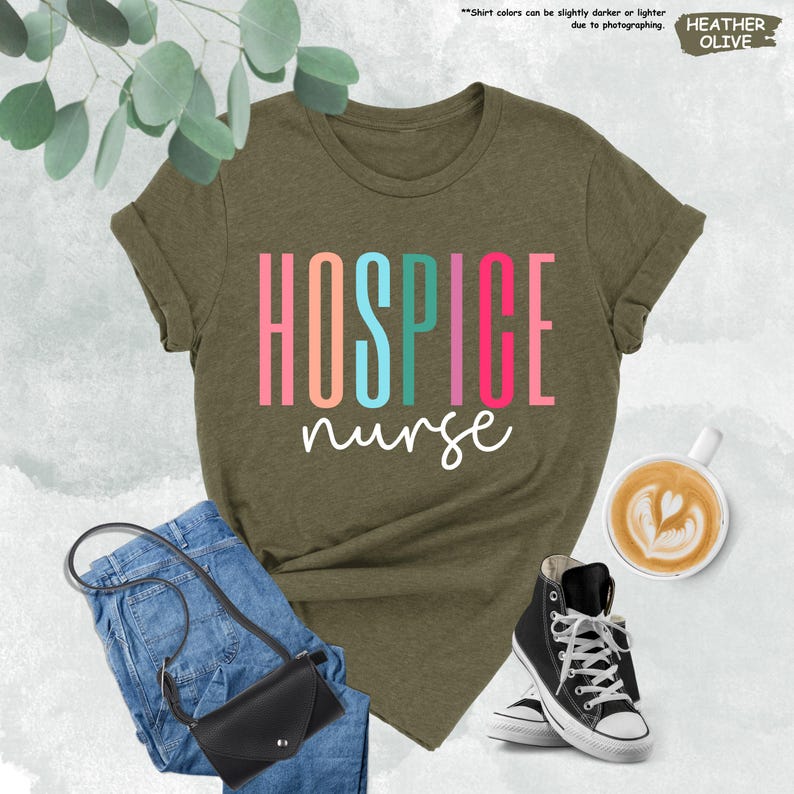 K&ouml;nnte beinhalten: Ein gr&uuml;nes T-Shirt mit dem Text "HOSPICE nurse" in bunten Buchstaben. Das Shirt liegt auf einer wei&szlig;en Oberfl&auml;che mit einer Jeans, einer Handtasche und einer Tasse Kaffee.