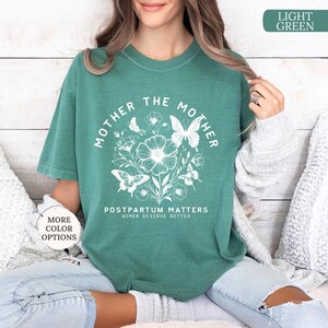 Może przedstawiać: Jasnozielony t-shirt z białym nadrukiem graficznym przedstawiającym tekst "Mother the Mother" w k&oacute;łku z motylami i kwiatami. Tekst "Postpartum Matters Women Deserve Better" jest nadrukowany pod k&oacute;łkiem.