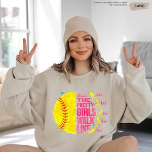 May include: Sand-colored sweatshirt with a softball graphic. The graphic features a yellow softball with red stitching and the text "ALL THE PRETTY GIRLS WALK LIKE THIS!" in pink and white. The sweatshirt is a casual style.