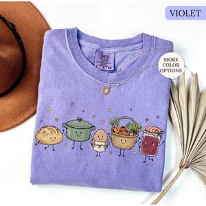 May include: A violet t-shirt featuring a graphic print of a smiling loaf of bread, a green pot, an egg wearing an apron, a basket of vegetables, and a jar of jam. The shirt has a crew neck and a Comfort Colors label on the inside collar.
