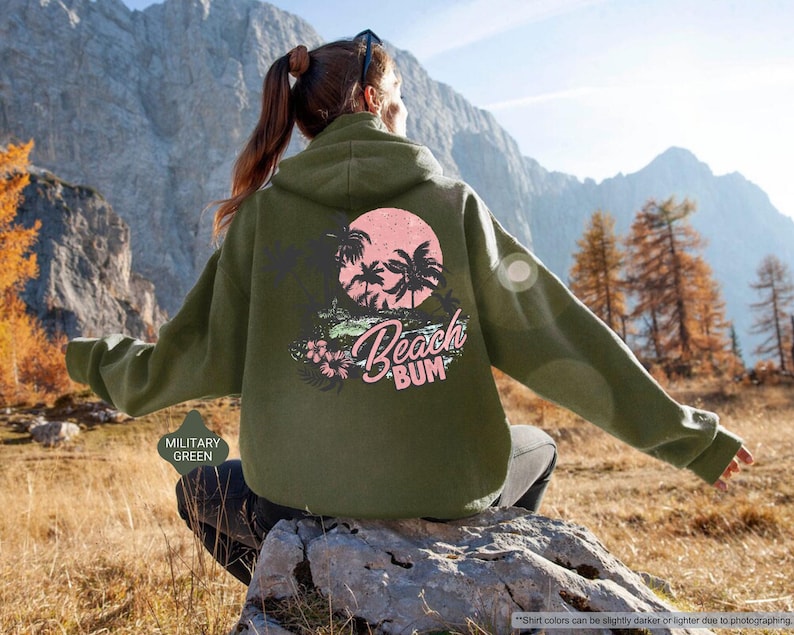 May include: Military green hoodie with a graphic on the back showing a pink sun, palm trees, and the words "Beach Bum." The hoodie has a relaxed fit and a drawstring hood.
