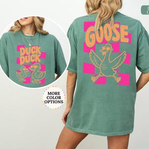 May include: A light green t-shirt featuring a back graphic of a goose wearing sunglasses above the text GOOSE, set against a pink checkered pattern. The front chest graphic displays two ducks wearing sunglasses with the text DUCK DUCK.
