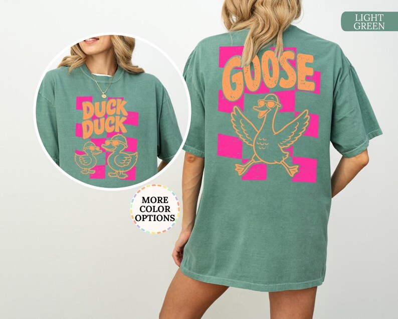May include: A light green t-shirt featuring a back graphic of a goose wearing sunglasses above the text GOOSE, set against a pink checkered pattern. The front chest graphic displays two ducks wearing sunglasses with the text DUCK DUCK.
