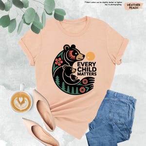 May include: Heather peach-colored t-shirt with a graphic of a bear family and the text "EVERY CHILD MATTERS". The design includes a flower, trees, and a sun. The shirt is paired with blue jeans and beige flats.