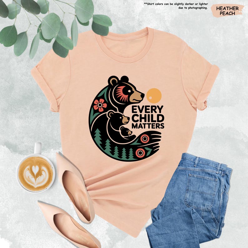 May include: Heather peach-colored t-shirt with a graphic of a bear family and the text "EVERY CHILD MATTERS". The design includes a flower, trees, and a sun. The shirt is paired with blue jeans and beige flats.