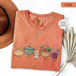 May include: A burnt orange crewneck t-shirt featuring a graphic print of a smiling loaf of bread, a green pot, an egg in an apron, a basket of vegetables, and a jar of jam. The shirt has a Comfort Colors label and small colorful stars and hearts.