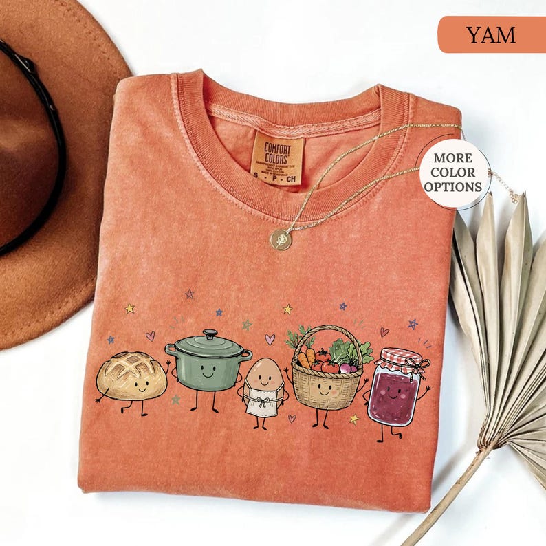May include: A burnt orange crewneck t-shirt featuring a graphic print of a smiling loaf of bread, a green pot, an egg in an apron, a basket of vegetables, and a jar of jam. The shirt has a Comfort Colors label and small colorful stars and hearts.