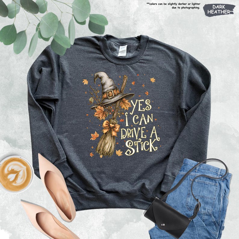 May include: A dark heather gray crewneck sweatshirt featuring a graphic of a witch hat, a broomstick with a bow, and scattered autumn leaves. The text reads, "YES I CAN DRIVE A STICK." The design includes orange, brown, and tan colors on the fabric.