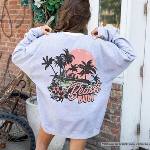May include: A light grey sweatshirt with a beach-themed graphic on the back. The design features black palm trees, a pink sunset, and the words "Beach Bum" in pink and white script. The sweatshirt is a casual style.