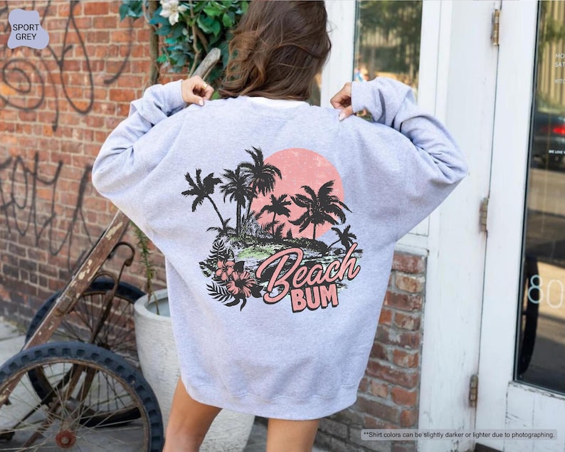 May include: A light grey sweatshirt with a beach-themed graphic on the back. The design features black palm trees, a pink sunset, and the words "Beach Bum" in pink and white script. The sweatshirt is a casual style.