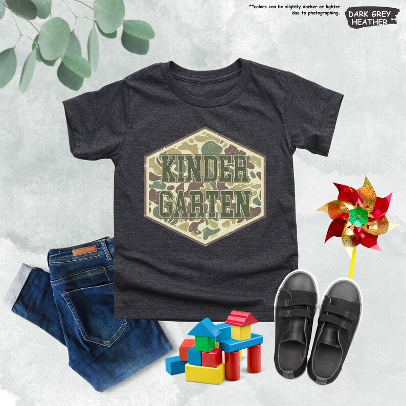 May include: Dark grey heather t-shirt with a camouflage design that reads "KINDER GARTEN". The image also includes a pair of dark blue jeans, black trainers, a colourful pinwheel, and wooden building blocks.