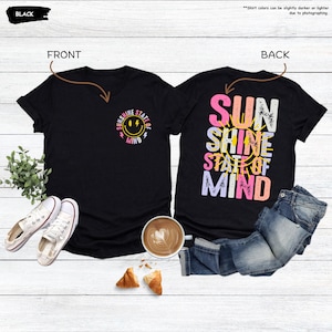 Pu&ograve; includere: T-shirt nero con scollo a V con grafica frontale di una faccia sorridente e le parole "Sunshine State of Mind". La parte posteriore della maglietta presenta le parole "Sun Shine State of Mind" in un design colorato e stratificato. Sono visibili anche scarpe da ginnastica bianche e jeans.