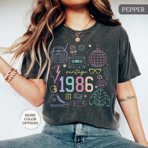 May include: Dark gray t-shirt with a colorful "vintage 1986" graphic. The design includes a boombox, cassette tape, disco ball, and other 80s-themed icons. The shirt has a crew neck and short sleeves.