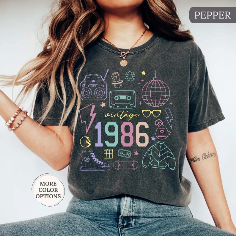 May include: Dark gray t-shirt with a colorful "vintage 1986" graphic. The design includes a boombox, cassette tape, disco ball, and other 80s-themed icons. The shirt has a crew neck and short sleeves.