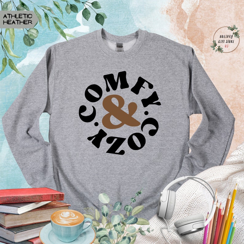 以下が含まれることがあります： グレーのトレーナーに「Comfy & Cozy」という文字が円形にデザインされています。文字は黒で、アンパサンドは茶色です。