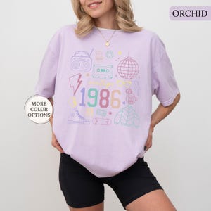 May include: Orchid-colored t-shirt with a vintage 1986 graphic. The design includes a boombox, cassette tape, disco ball, and other 80s-themed icons. The shirt is a relaxed fit and has short sleeves. The text "MORE COLOR OPTIONS" is on the left side of the image.