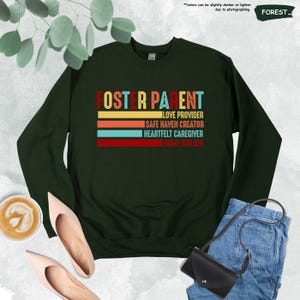 May include: A dark green sweatshirt with the text "Foster Parent" in colorful retro-style lettering. The text is stacked vertically and includes the phrases "Love Provider", "Safe Haven Creator", "Heartfelt Caregiver", and "Home Builder".