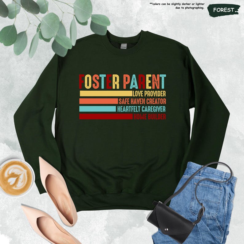 May include: A dark green sweatshirt with the text "Foster Parent" in colorful retro-style lettering. The text is stacked vertically and includes the phrases "Love Provider", "Safe Haven Creator", "Heartfelt Caregiver", and "Home Builder".
