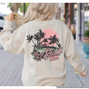 May include: Sand-coloured sweatshirt featuring a tropical beach graphic on the back. The design includes palm trees, a pink sun, and the words "Beach Bum". The sweatshirt has long sleeves and a crew neck.
