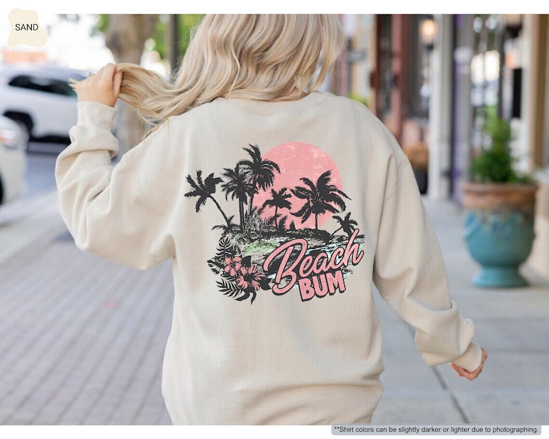 May include: Sand-coloured sweatshirt featuring a tropical beach graphic on the back. The design includes palm trees, a pink sun, and the words "Beach Bum". The sweatshirt has long sleeves and a crew neck.