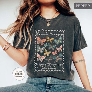 Puede incluir: Camiseta gris oscuro con un dise&ntilde;o de mariposas coloridas dentro de un marco blanco. El texto "Speech Therapy" y "helps little voices take flight" est&aacute; impreso. La camiseta es de estilo casual y c&oacute;modo.