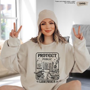 May include: A beige sweatshirt with a black graphic design that says "Protect Public Libraries". The graphic features a stack of books, a quill pen, and an inkwell.