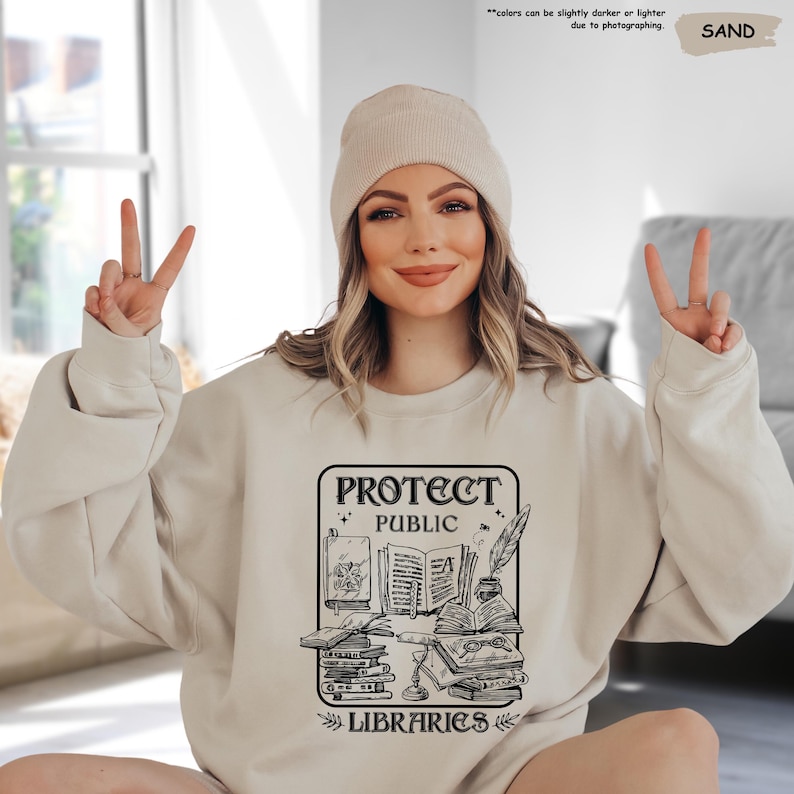 May include: A beige sweatshirt with a black graphic design that says "Protect Public Libraries". The graphic features a stack of books, a quill pen, and an inkwell.