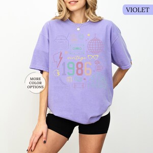 May include: A violet-colored t-shirt with a retro graphic design. The design includes a boombox, cassette tape, disco ball, and the year "1986" in colorful text. The shirt also has the text "vintage".