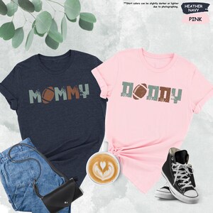 May include: Two t-shirts, one heather navy blue and one pink, with the words "MOMMY" and "DADDY" printed on them. Each shirt features a football graphic.