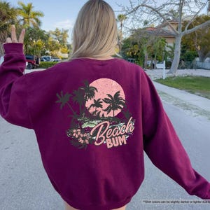May include: Maroon sweatshirt with a beach-themed graphic on the back. The design features palm trees, a pink sun, and the words "Beach Bum" in a script font. The sweatshirt is a solid maroon colour.