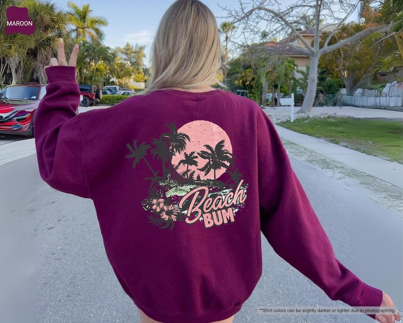 May include: Maroon sweatshirt with a beach-themed graphic on the back. The design features palm trees, a pink sun, and the words "Beach Bum" in a script font. The sweatshirt is a solid maroon colour.