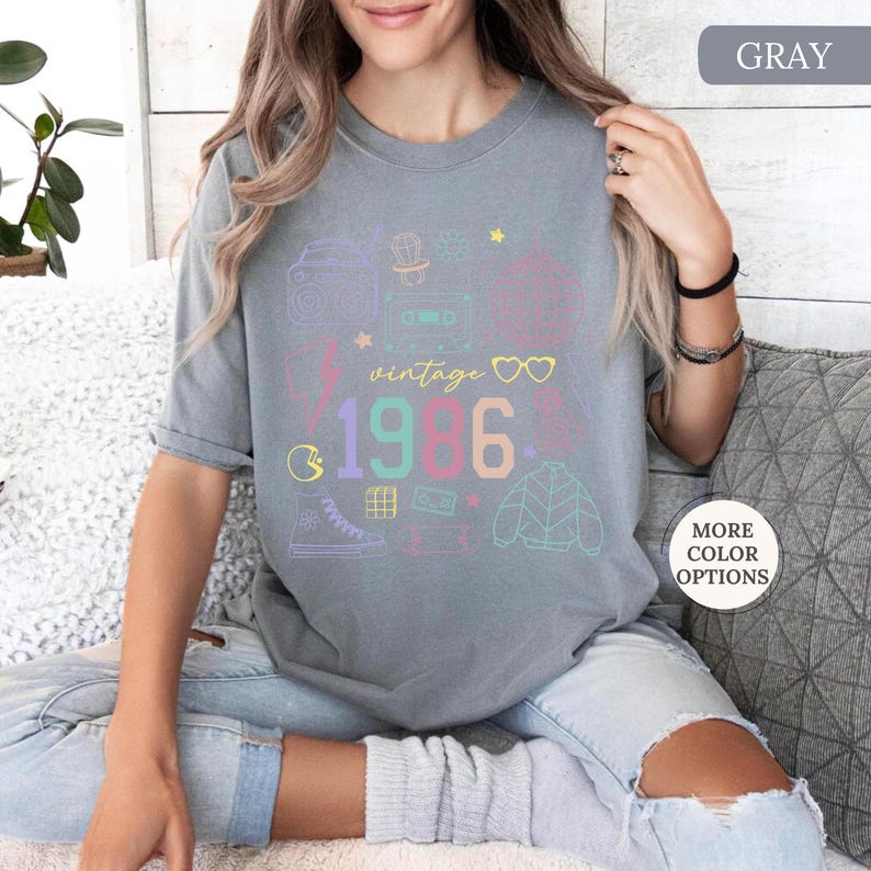 May include: Gray t-shirt with a colorful retro design. The design includes the word "vintage" above the numbers "1986", along with illustrations of a boombox, cassette tape, and other 80s-themed icons. The shirt is a comfortable, casual style.