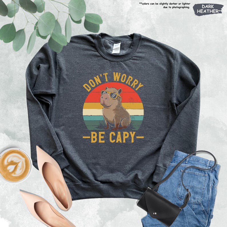 May include: Dark heather gray sweatshirt with a graphic of a capybara wearing sunglasses inside a retro sunset circle. Text reads "DON'T WORRY - BE CAPY-". The sweatshirt is paired with jeans, a black purse, and shoes.