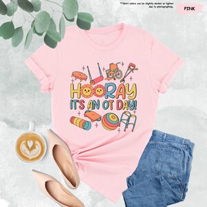 Peut inclure: T-shirt rose avec un motif graphique d'outils d'ergoth&eacute;rapie et le texte "HOORAY IT'S AN OT DAY!"